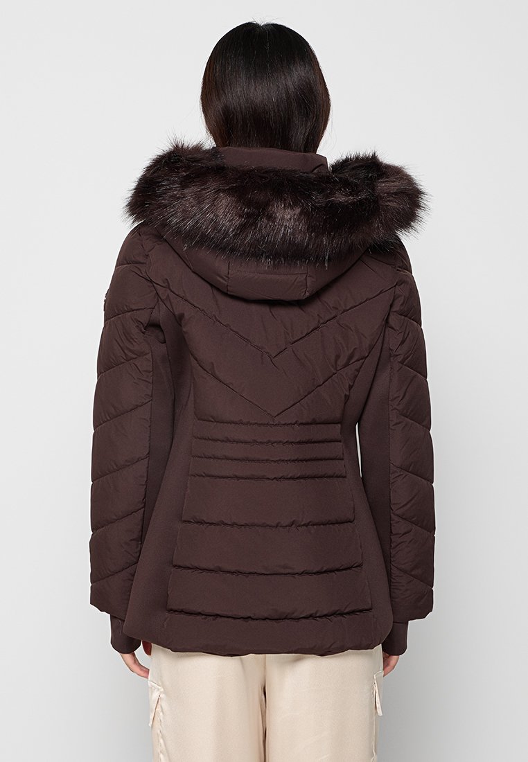 Vrouw in een donkerbruine gewatteerde jas met een met bont afgewerkte capuchon, staande met haar rug naar de camera tegen een effen achtergrond.