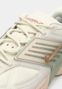 Közelkép egy bézs Nike sneaker cipőről, hálós anyaggal, vastag fűzőkkel, műanyag oldalsó panellel és narancssárga Nike logóval.