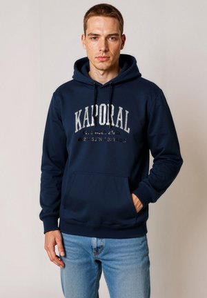 Sudadera azul marino de tejido suave con un bolsillo frontal tipo canguro, que presenta el texto "KAPORAL" en gris claro en el pecho.