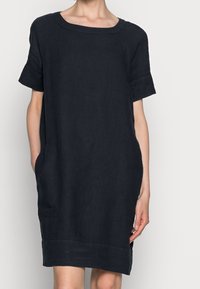 Žena v volném černém šatech s krátkým rukávem a čtvercovým výstřihem, s rukama v kapsách, na jednobarevném pozadí.