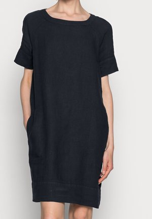 Femme portant une robe noire ample à manches courtes et décolleté carré, les mains dans les poches latérales, sur un fond uni.