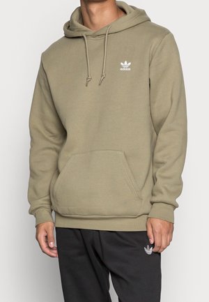 Mężczyzna w oliwkowej bluzie Adidas z kapturem i przednią kieszenią oraz czarnych spodniach Adidas na jednolitym tle.