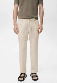 Beige linnen broeken met een relaxte pasvorm, een trekkoord in de taille en rechte pijpen, gecombineerd met zwarte sandalen en een gestructureerd olijfgroen shirt.