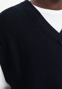 Schwarzer gestrickter ärmelloser Pullover mit tiefem V-Ausschnitt, der ein strukturiertes Muster und gerippte Kanten aufweist, über einem weißen Hemd getragen.
