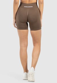 Modell in braunen Sportshorts mit dem Markennamen "SMILODOX" auf dem Bund, stehend vor einem neutralen Hintergrund.