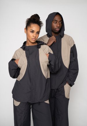 A man and woman wearing matching black and beige hooded jackets and pants, standing closely against a plain background.