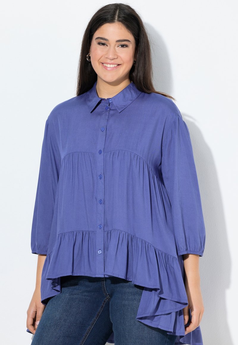 Blusa viola con colletto, caratterizzata da dettagli arricciati e un orlo posteriore più lungo. Realizzata in tessuto leggero, con maniche a palloncino.