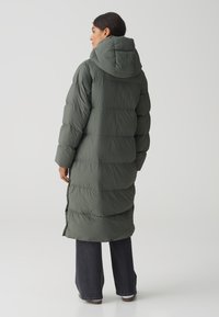 Donkergroene pufferjas met een capuchon, met een gequilt ontwerp en een knielange snit. Gedragen over donkere jeans en bruine schoenen.