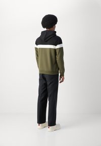 Hettegenser i olivengrønn med en svart panel og hvit stripe, kombinert med svarte bukser og kremfargede joggesko. Glatt tekstur.