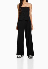 Top nero senza spalline con chiusura frontale a bottoni, abbinato a pantaloni neri larghi e sneakers grigie sportive. Tessuto dalla texture liscia.