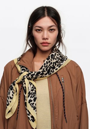 Mujer joven con cabello largo y oscuro lleva una chaqueta marrón, camiseta amarillo claro y una bufanda con estampado de leopardo atada alrededor del cuello sobre un fondo blanco.