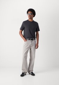 T-shirt en coton noir, jean denim gris clair, ceinture noire et baskets noires avec des accents blancs. Le mannequin porte des lunettes de soleil et a une coiffure afro.