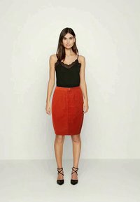 Mulher vestindo um top de renda preta, uma saia em tom de ferrugem até os joelhos e sapatos de salto preto com tiras no tornozelo, de pé contra um fundo simples.