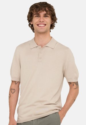 Jeune homme aux cheveux bouclés souriant, portant un polo beige à manches courtes et montrant des tatouages sur les deux avant-bras, debout avec une main dans la poche.