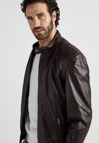 Brun läderjacka med dragkedjestängning, knappkrage och sidoficka. Slät textur, skräddarsydd passform och subtila sömsdetaljer.