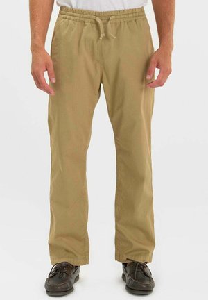 Uomo che indossa pantaloni beige con coulisse e scarpe casual marrone scuro, in piedi con le mani rilassate ai lati.