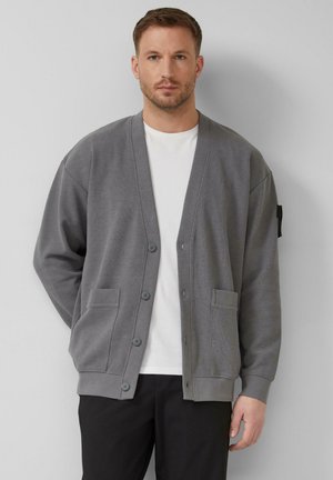 Mann trägt eine graue, geknöpfte Strickjacke über einem weißen T-Shirt und schwarzen Hosen, steht mit verschränkten Händen hinter dem Rücken vor einem einfarbigen Hintergrund.