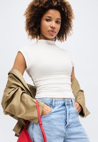 Γυναίκα με σγουρά μαλλιά που φοράει λευκή εφαρμοστή μπλούζα, ανοιχτό μπλε τζιν και χακί μπουφάν που έχει περασμένο στους βραχίονες της, κρατώντας μια κόκκινη τσάντα χειρός.