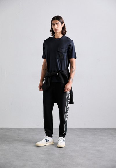 Bărbat tânăr cu p�ăr până la umeri, purtând un tricou negru, pantaloni de trening negri cu text alb pe lateral, adidași albi și un hanorac negru legat în jurul taliei.