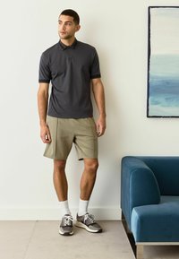Mørkegrå polo t-shirt med mønstret krave, parret med lyse khaki shorts. Modellen har hvide strømper og grå sneakers på, og står tæt på en blå sofa.