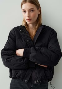 Jeune femme avec de longs cheveux blonds portant une veste matelassée noire avec fermeture éclair et boutons-pression, les bras croisés, sur un fond uni clair.