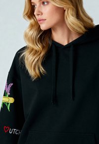 Sudadera negra con cordón, con coloridos bordados en lavanda y una venda. Incluye un bolsillo frontal y una textura suave.