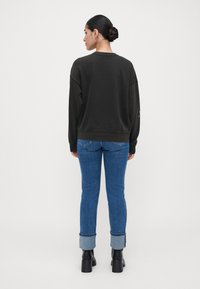 Kvinne med hår i knute iført svart hettegenser, oppbrettede blå jeans og svarte ankelstøvler, som står med ryggen til foran en enkel hvit bakgrunn.