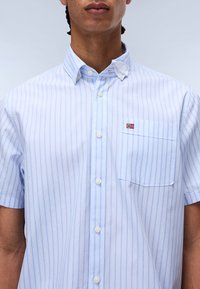 Chemise à manches courtes bleue à rayures en coton, dotée d'un col boutonné, d'une poche à l'avant et d'un petit emblème du drapeau norvégien.