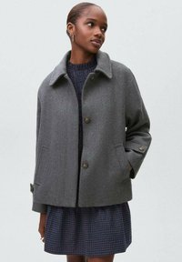 Veste en mélange de laine grise avec un col, fermeture à boutons à l'avant et poches latérales ; portée par-dessus une robe à carreaux bleu marine. Texture lisse, coupe décontractée.