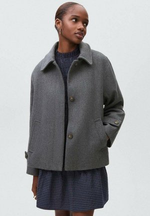 Veste en mélange de laine grise avec un col, fermeture à boutons à l'avant et poches latérales ; portée par-dessus une robe à carreaux bleu marine. Texture lisse, coupe décontractée.