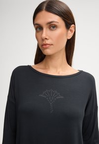 Czarna bluzka z długim rękawem wykonana z gładkiej tkaniny, ozdobiona dekoracyjnym wzorem z kryształków przypominającym liść na środku.
