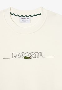 Felpa color crema con colletto a girocollo, caratterizzata da testo e logo "LACOSTE" ricamati in verde. Tessuto morbido e strutturato con una vestibilità ampia.