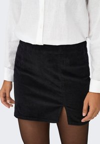 Mini-jupe noire en velours côtelé avec une fente à l'avant, portée avec des collants noirs transparents et une chemise blanche boutonnée à manches longues.