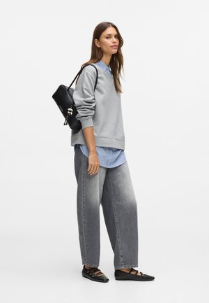 Kobieta stoi w szarym sweatshirtcie na niebieskiej koszuli w kratę, w szerokich szarych dżinsach, czarnych balerinkach i z czarną torbą na ramię ozdobioną breloczkiem.