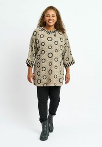 Lange schwarze Tunika mit schwarzen Kreis-Designs, gestreiften Manschetten, kombiniert mit schwarzen Hosen und schwarzen Knöchelstiefeln. Strukturierter Stoff.