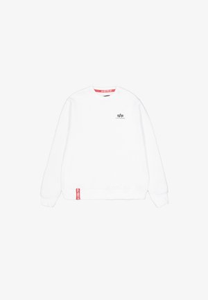 Biała, długa bluza z okrągłym dekoltem i długim rękawem z małym czarnym logo Alpha Industries na lewej piersi oraz czerwoną metką blisko dolnego brzegu.