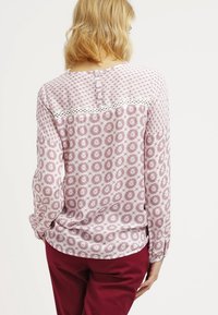 Blusa a maniche lunghe rosa chiaro con motivi circolari, dettagli in pizzo al giogo e chiusura con bottoni sul retro. Tessuto morbido e leggero.