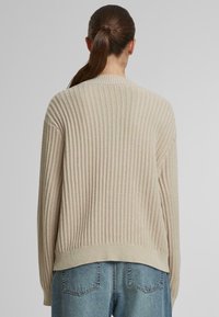 Pulover din tricot bej, cu o siluetă confortabilă, având guler rotund și un fit ușor supradimensionat, asortat cu blugi din denim de culoare albastru deschis.