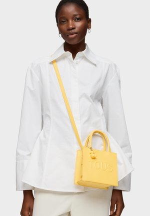 Mujer con camisa blanca de botones y pantalones color crema, llevando un pequeño bolso bandolera amarillo con el grabado "TOUS".