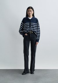 Femme debout portant un pull rayé bleu marine avec des boutons, un pantalon noir avec de grandes poches avant, des chaussures noires et des boucles d'oreilles créoles dorées.