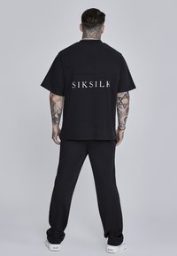 Crni pamučni majica s SIKSILK logom na leđima, u kombinaciji s odgovarajućim crnim hlačama i bijelim tenisicama. Glatka tekstura i opušteni kroj.