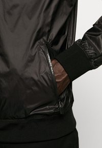 Czarna kurtka typu bomber o gładkim, błyszczącym wykończeniu, wyposażona w zapinaną na zamek kieszeń boczną, elastyczne mankiety oraz prążkowany pas biodrowy.