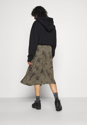 Woman with curly hair wearing a black hoodie, patterned midi skirt, gray socks, and black sneakers, standing against a plain white background.