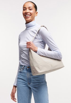 Frau trägt hellblauen Rollkragenpullover und Jeans, lächelt und hält eine beige Schultertasche vor einem schlichten weißen Hintergrund.