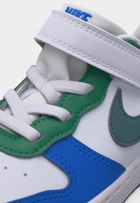 Gros plan sur une chaussure de sport Nike blanche avec des panneaux en cuir vert et bleu, des lacets blancs et une bride blanche sur le dessus.