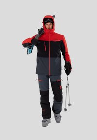 Skijakke i rød og sort, med en lynlåslomme på brystet og en justerbar hætte. Parret med matchende bukser og grå skistøvler.