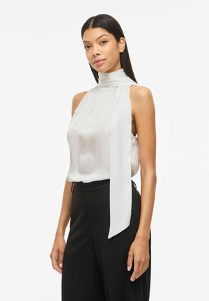 Femme aux longs cheveux foncés portant une blouse blanche sans manches à col montant avec un long ruban en tissu et un pantalon noir taille haute.