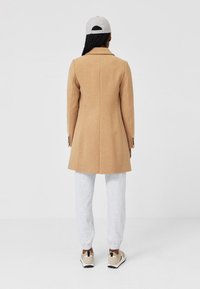 Cappotto di lana color sabbia con un design aderente, davanti a bottone singolo e lapels sulle spalle. Abbinato a pantaloni della tuta grigi e sneakers beige.