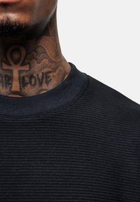 Schwarzer, strukturiert Pullover mit einem gerippten Ausschnitt, im Hintergrund ist eine Nahaufnahme von kunstvollen Nackentattoos zu sehen.