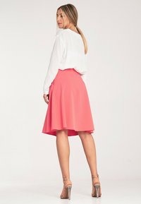 Koralowa spódnica w kształcie dzwonu o gładkiej fakturze, zestawiona z białą bluzką z długim rękawem. Wyraźny detal wysokich sandałów na obcasie z paskami na kostkach.